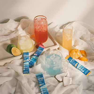 Four colorful drinks with fruit and supplement packets on a white cloth background.