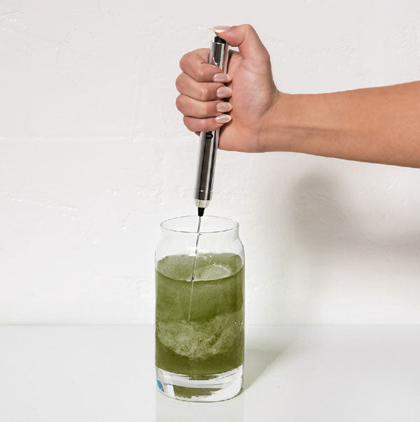 Hand stirring green liquid in a glass with a metal frother.
